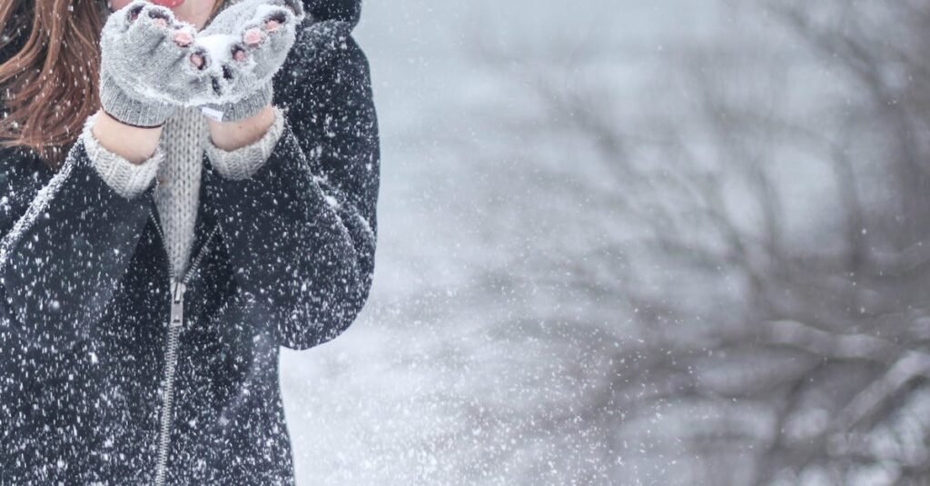 Woman in cozy winter clothing blowing snowflakes with excitement outdoors in a snowy setting.
