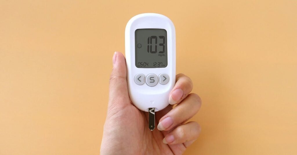 A hand holding a blood glucose meter displaying 103 mg/dL on a neutral background.